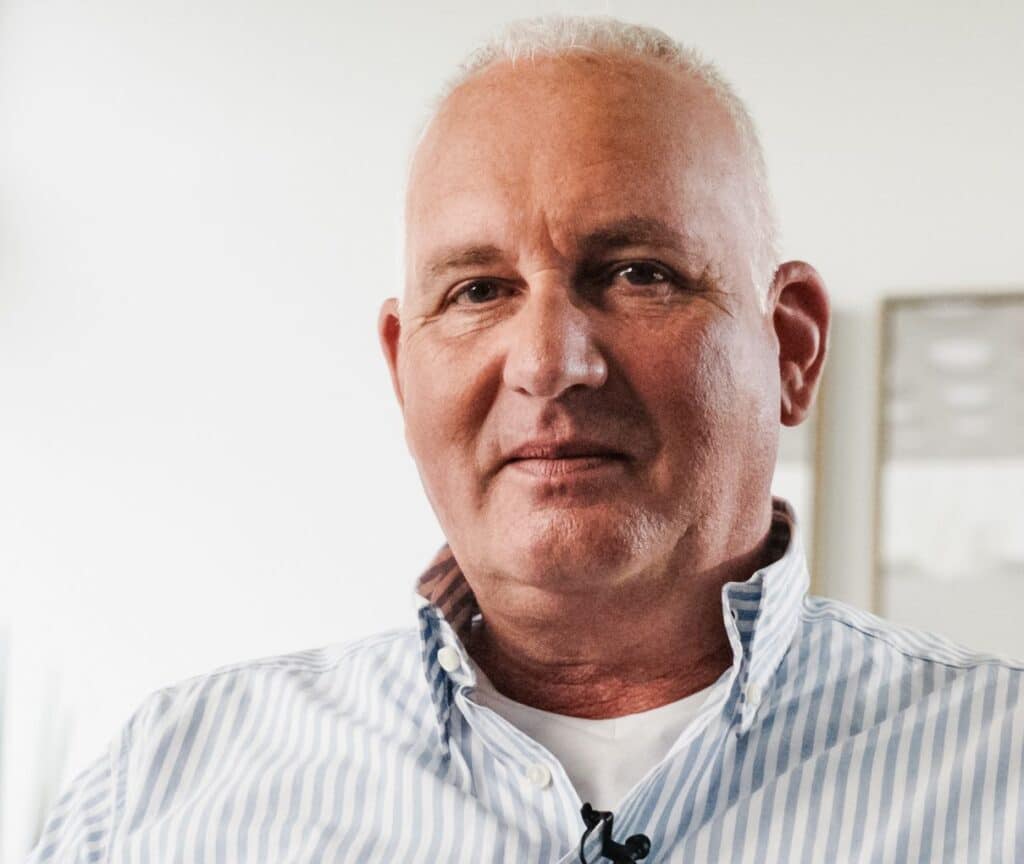 Ein Mann mit kurzen weißen Haaren, der ein gestreiftes Hemd und ein weißes T-Shirt trägt, blickt in die Kamera, während im Hintergrund Bauarbeiter mit Straßenbauarbeiten beschäftigt sind.