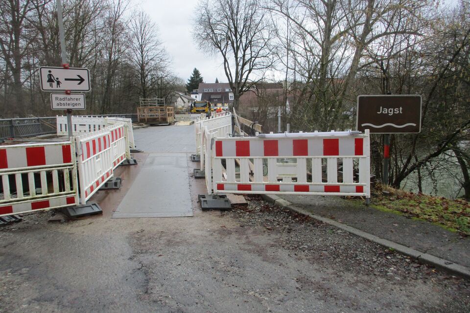 Ein mit Barrieren blockierter Weg führt zu einer im Bau befindlichen Brücke. Im Hintergrund sind Wegweiser und Bäume zu sehen. Dieses Hochbauprojekt legt Wert auf nachhaltige Baupraktiken und Recyclingmethoden, um die Umweltbelastung zu minimieren.