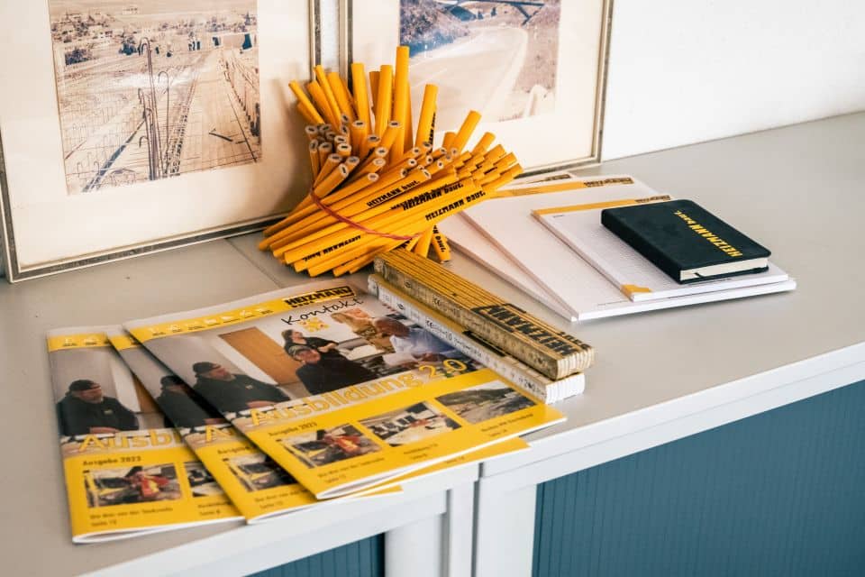 Schreibtisch mit gelben Stiften, Linealen, Veranstaltungsflyern, Notizbüchern und gerahmten Fotos an der Wand im Hintergrund – alles Materialien, die sich leicht für das Recycling organisieren lassen.