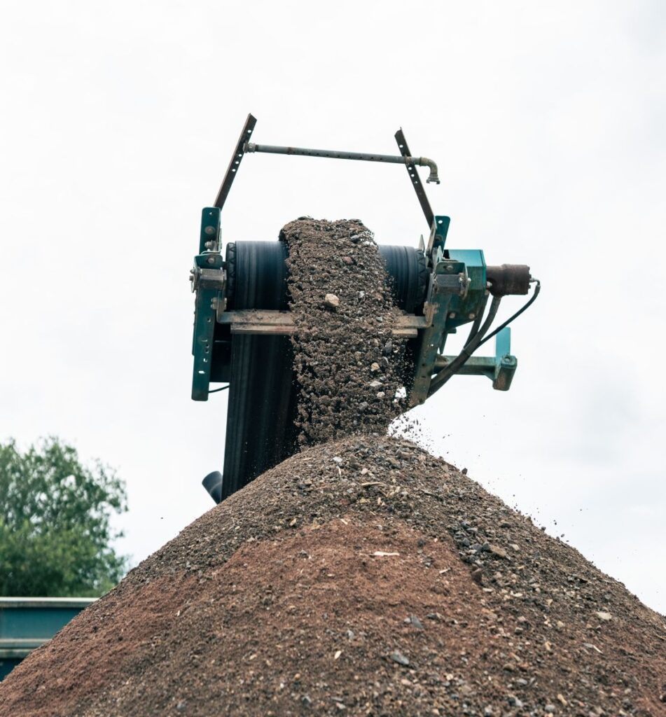 Ein Förderband befördert einen Erd- oder Kieshaufen mit sichtbaren mechanischen Teilen vor dem Hintergrund eines wolkigen Himmels ab und verdeutlicht so die robuste Effizienz der Bau-Techniken.