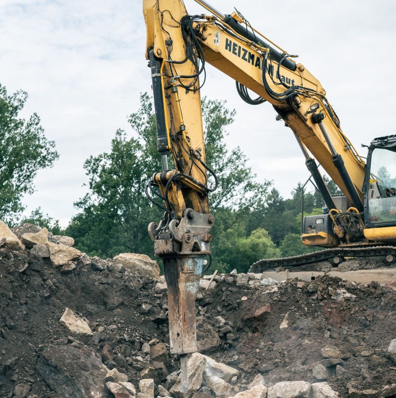 Ein gelber Bagger mit einem hydraulischen Brecheraufsatz bricht auf einer Baustelle Steine und bereitet so effizient den Boden für Straßenbauprojekte vor.