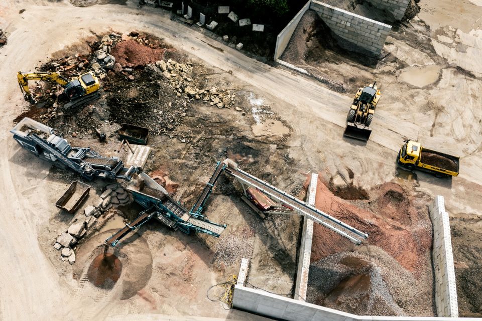 Luftaufnahme einer Baustelle mit schweren Maschinen, darunter Bagger und Bulldozer, die Steine und Kies sortieren. Diese dynamische Szene zeigt Hochbau- und Straßenbauarbeiten im Gange.