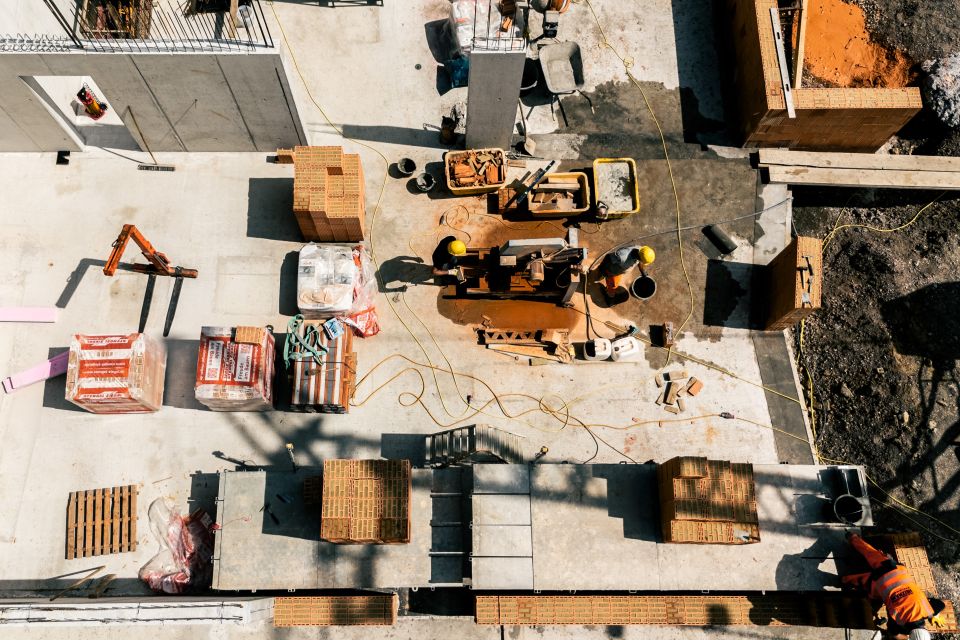 Luftaufnahme einer Baustelle mit Arbeitern, Ziegeln, Maschinen und überall verstreuten Baumaterialien. Die Szene zeigt Elemente von Tiefbau- und Hochbauprojekten im Gange.