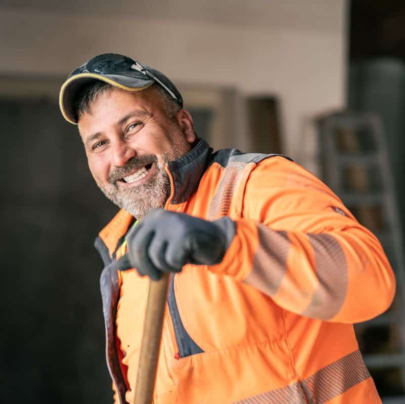 Ein lächelnder Arbeiter in einer leuchtend orangefarbenen Jacke und Mütze steht mit einem Werkzeug in der Hand in einer Inneneinrichtung einer Baufirma.