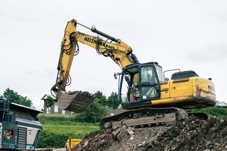Ein gelber Bagger auf einer Hochbau-Baustelle hebt einen Eimer Erde an, im Hintergrund sind Grünflächen und ein bewölkter Himmel zu sehen.