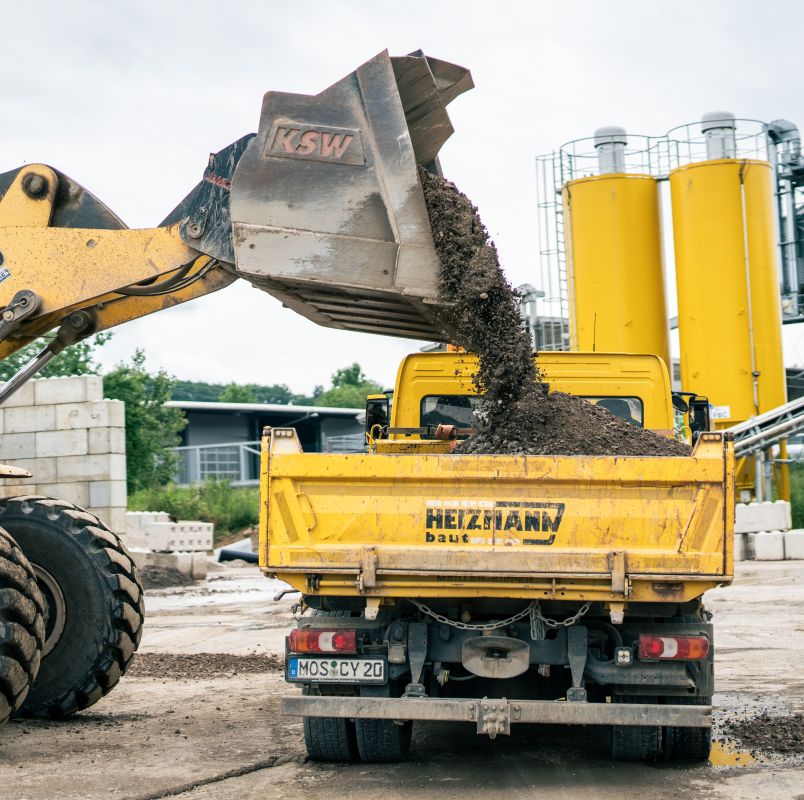 Eine Baustelle mit einem gelben Lastwagen, der von einem Frontlader mit Kies beladen wird. Im Hintergrund sind Industrietanks zu sehen, die die Rolle des Recyclings in modernen Bauprojekten hervorheben.