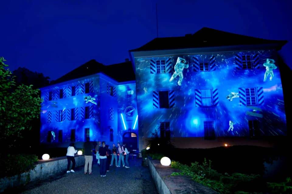 Ein Gebäude, das nachts mit blauen Lichtern und weltraumbezogenen Projektionen geschmückt ist und vor dem Menschen stehen, zeigt die innovativen Aspekte von Hochbau.