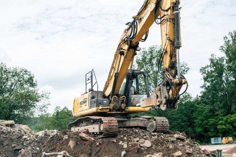 Gelber Bagger auf einem Haufen aus Erde und Steinen, entscheidend für Hochbauprojekte, mit Bäumen im Hintergrund unter einem bewölkten Himmel.