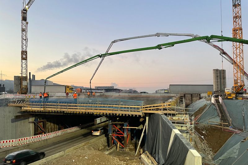 Auf der geschäftigen Baustelle sind fleißige Arbeiter, riesige Kräne und eine Betonpumpe über der Fahrbahn im Einsatz, alles vor dem Hintergrund des frühen Morgen- oder Abendhimmels – ein wahrer Beweis für den modernen Hochbau.