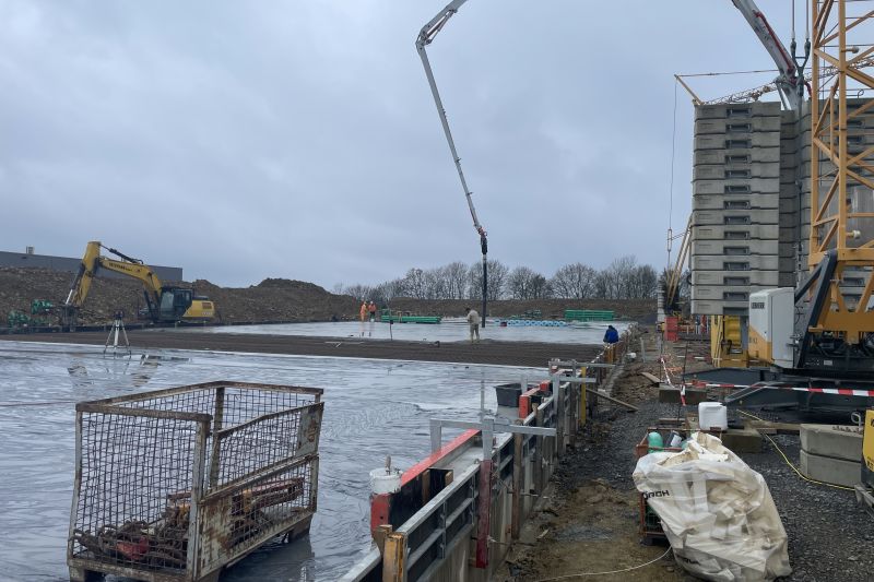 Baustelle mit Kränen, Baggern und Arbeitern, die unter bewölktem Himmel das Fundament legen.