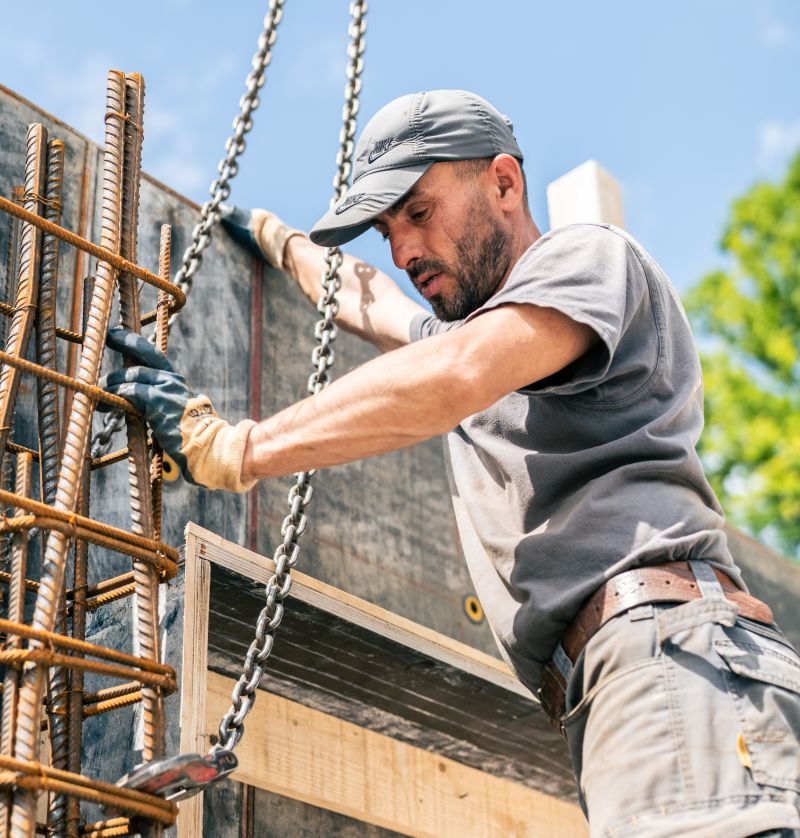 Ein Bauarbeiter mit Mütze und Handschuhen richtet an einem klaren Tag auf einer Baustelle Bewehrungsstäbe mit einer Kette aus und stellt so sein Fachwissen sowohl im Hoch- als auch im Straßenbau unter Beweis.