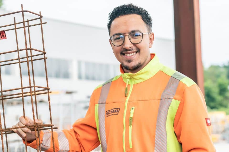Lächelnder Bauarbeiter in einer orangefarbenen Sicherheitsweste hält ein Metallgerüst auf einer Straßenbau-Baustelle.