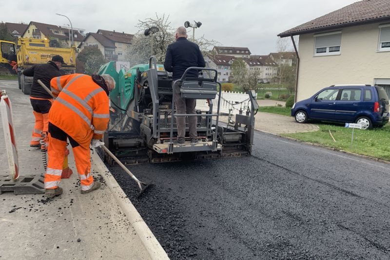 Arbeiter in orangefarbener Schutzkleidung eines örtlichen Bauunternehmens bedienen Maschinen, um in einer Wohnstraße in der Nähe von Häusern und geparkten Autos neuen Asphalt zu verlegen, und zeigen so ihr Engagement für das Recycling von Materialien.